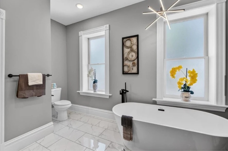 Freestanding bathtub under tall white-trimmed windows with gray walls and glossy tiled flooring