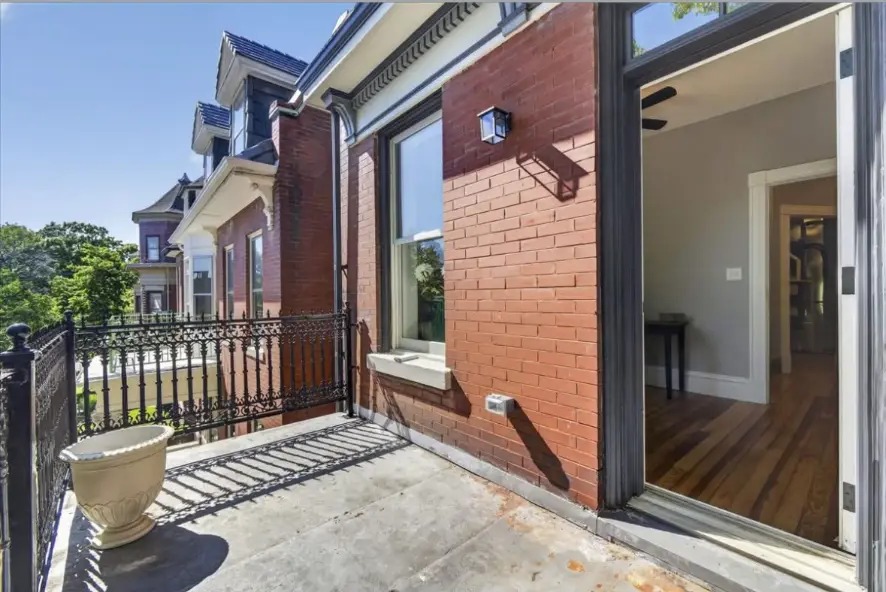 Balcony with black iron railing beside a red brick wall and open glass door
