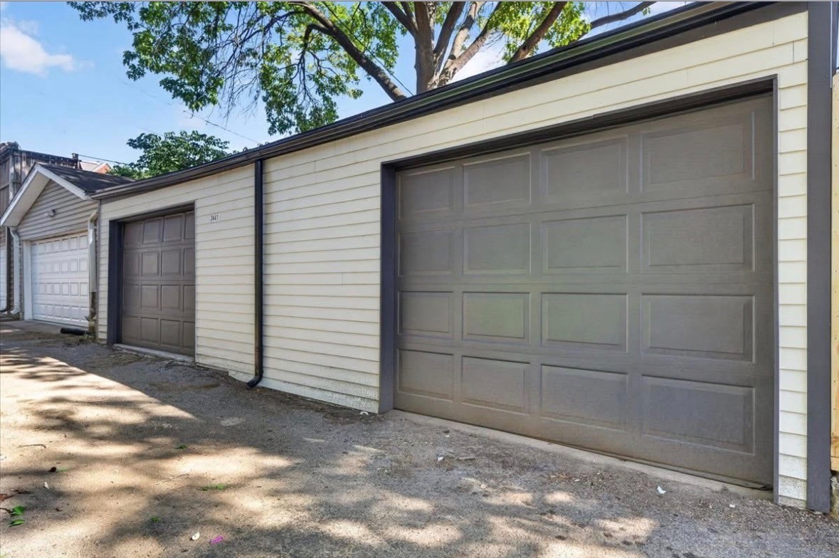 Detached garages with paneled doors, light siding and dark trim along a gravel drive