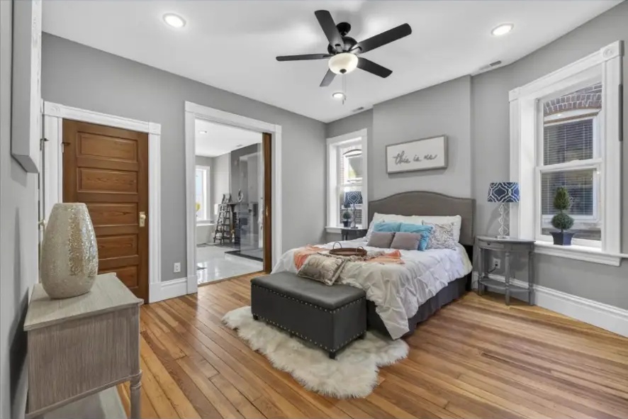 Gray bedroom with tall trimmed windows, hardwood floors, upholstered bed and ceiling fan