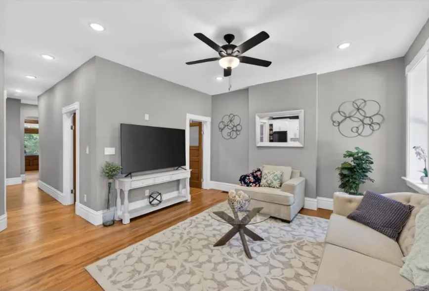 Living room with television console, ceiling fan, pale seating and hardwood flooring
